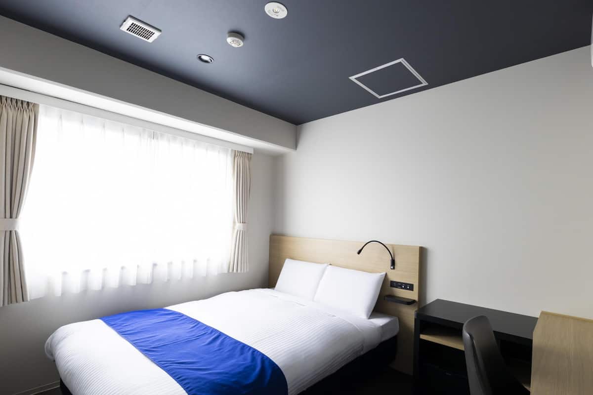 A modern hotel room with a neatly made bed, a desk with a chair, and a large window with curtains.