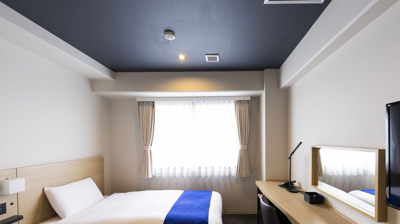 Quarto de hotel com cama, escrivaninha, luminária e janela com cortinas.