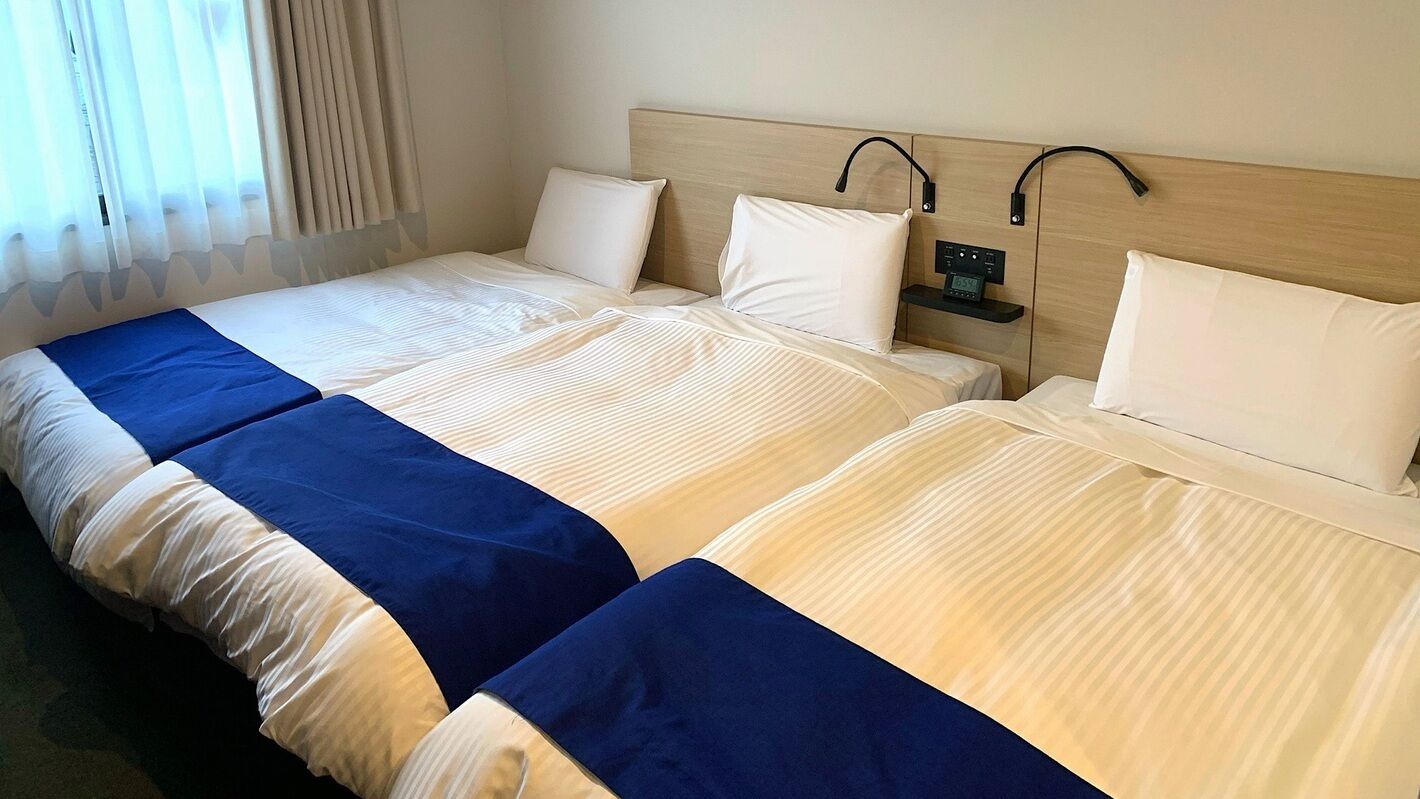 Quarto de hotel com duas camas, cada uma com cabeceira, mesa de cabeceira com relógio e janela com cortinas.