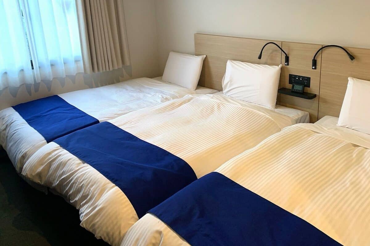 A hotel room with two beds, each with a headboard, a bedside table with a clock, and a window with curtains.