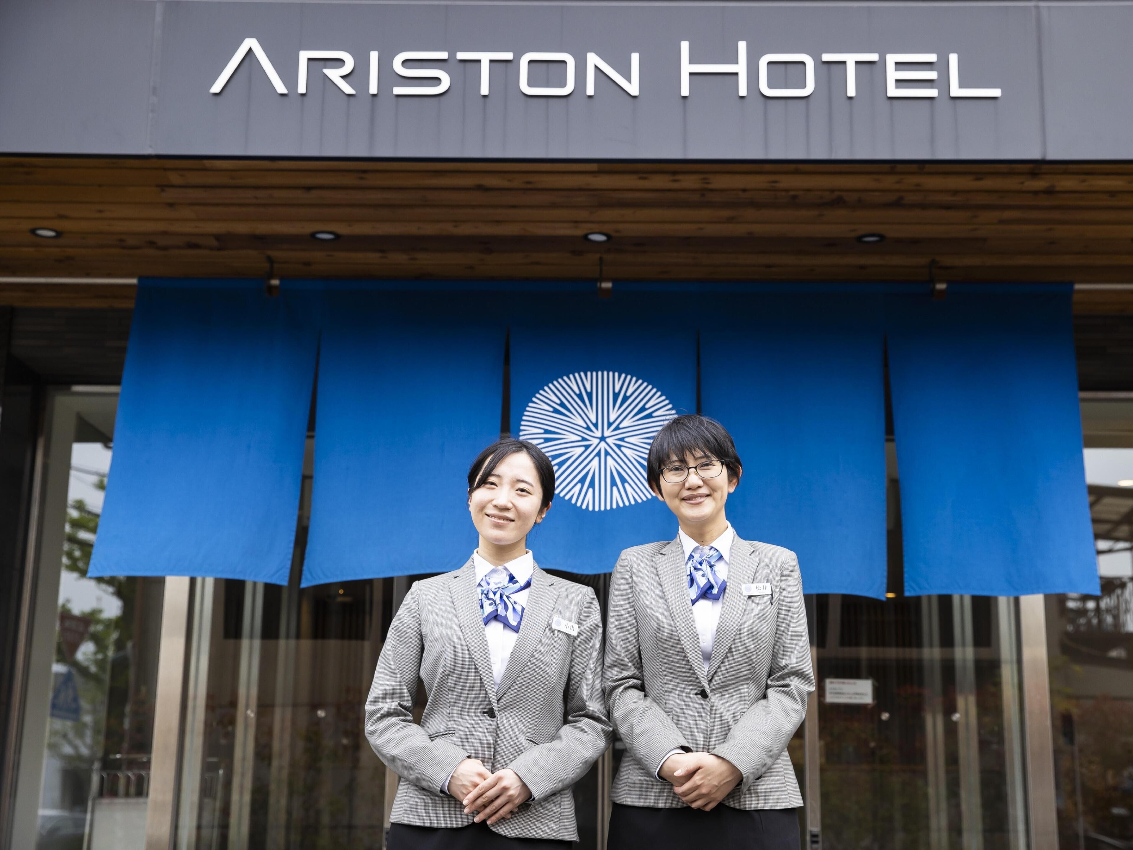 Two individuals in business attire standing in front of the Ariston Hotel entrance.