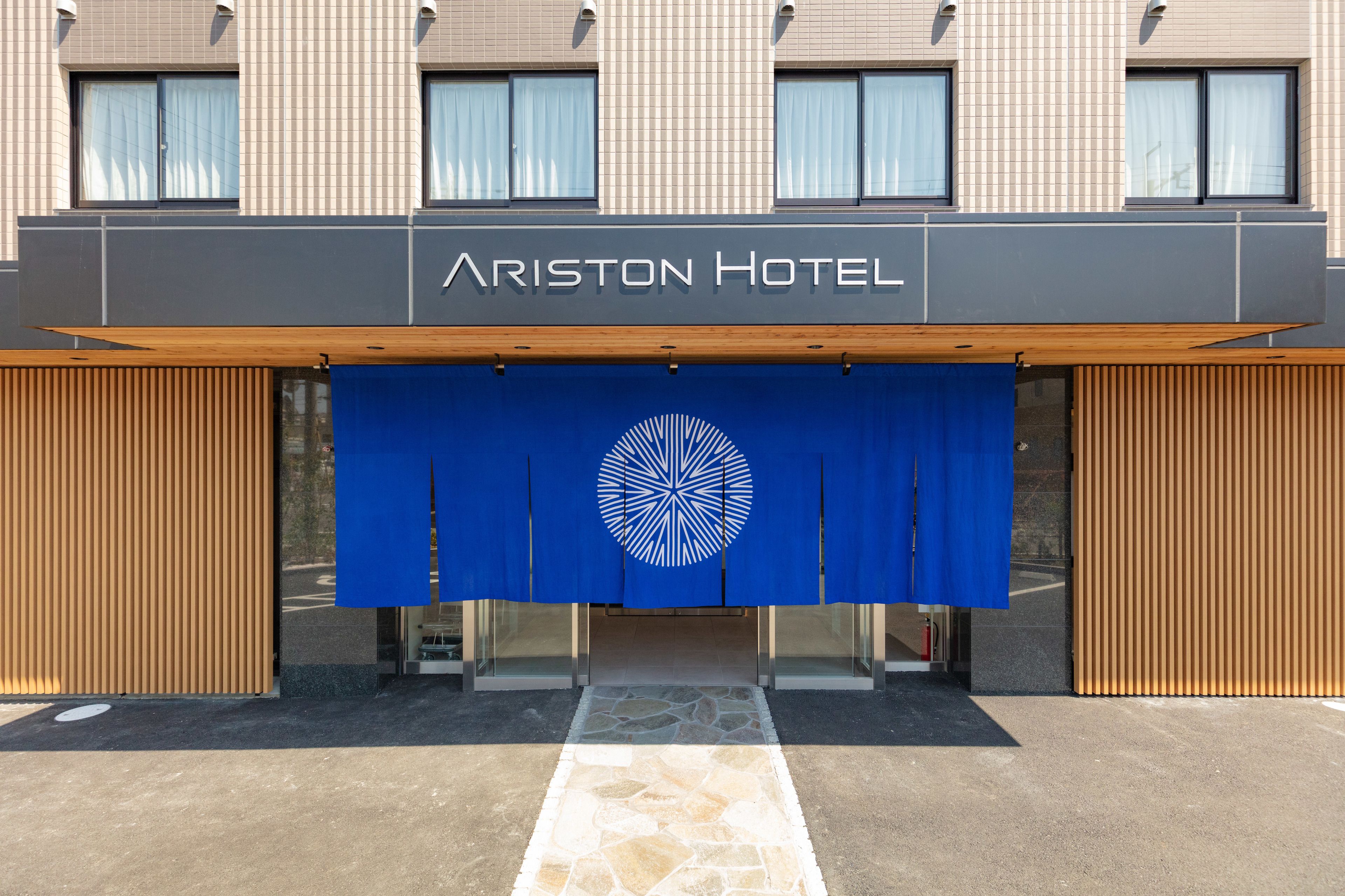 The exterior of the Ariston Hotel with a modern facade, a prominent entrance, and a blue awning with a circular logo.