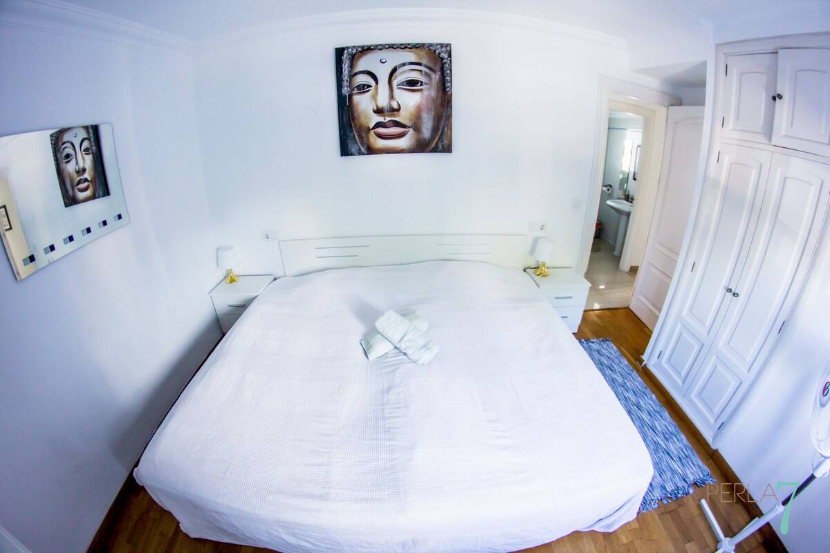 Een net opgemaakt bed met een witte deken en een opgevouwen handdoek, een witte nachtkastje met een lamp, en een aan de muur hangend schilderij.