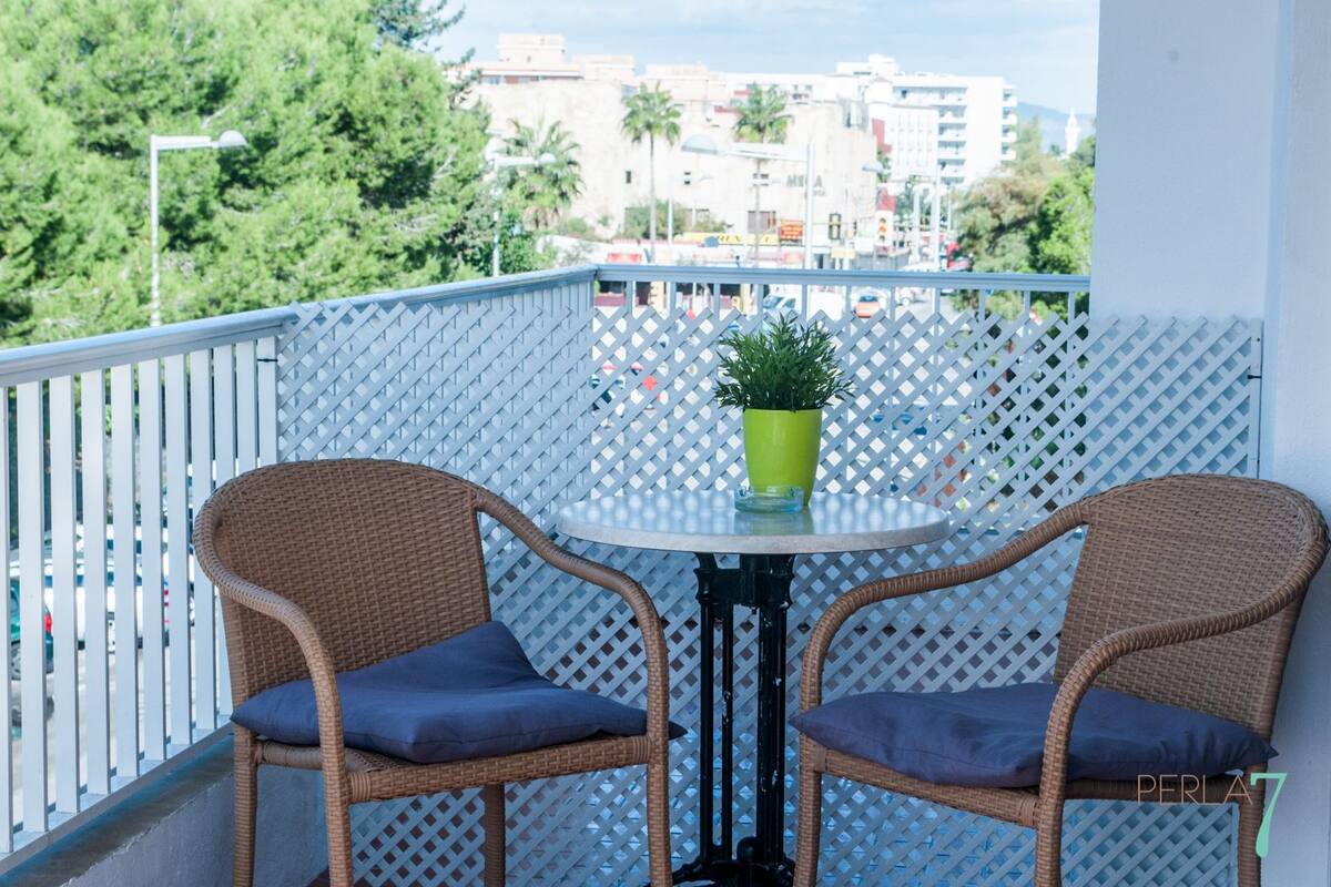 Een balkon met twee rieten stoelen en een kleine tafel, een potplant en uitzicht op een stadsgezicht met gebouwen en bomen.