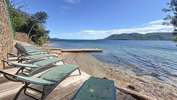 A wooden deck with lounge chairs facing a calm sea, a stone wall, and a clear sky. - Les Pavillons du Belvédère (Porto-Vecchio)