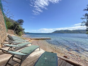 A wooden deck with lounge chairs facing a calm sea, a stone wall, and a clear sky. - Les Pavillons du Belvédère (Porto-Vecchio)