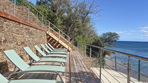 A wooden deck with lounge chairs overlooking a body of water, a stone wall, and a metal railing. - Les Pavillons du Belvédère (Porto-Vecchio)