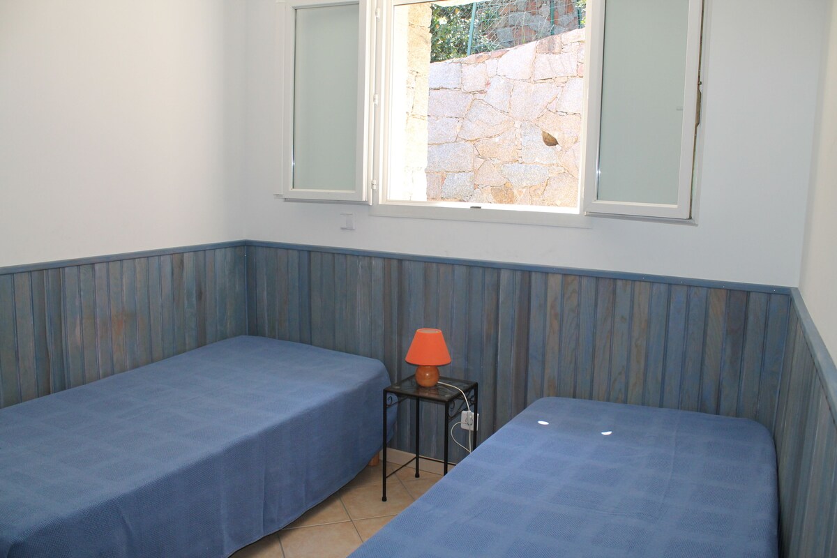 Una habitación con dos camas individuales, una mesita con una lámpara y una ventana que ofrece vistas a un muro de piedra y follaje exterior.