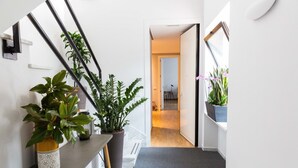 Un pasillo con una escalera, plantas en macetas, un banco gris y una puerta que da a otra habitación.