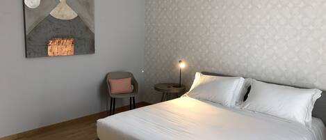 A neatly arranged bedroom with a large bed, a small side table, a chair, and a wall-mounted artwork.