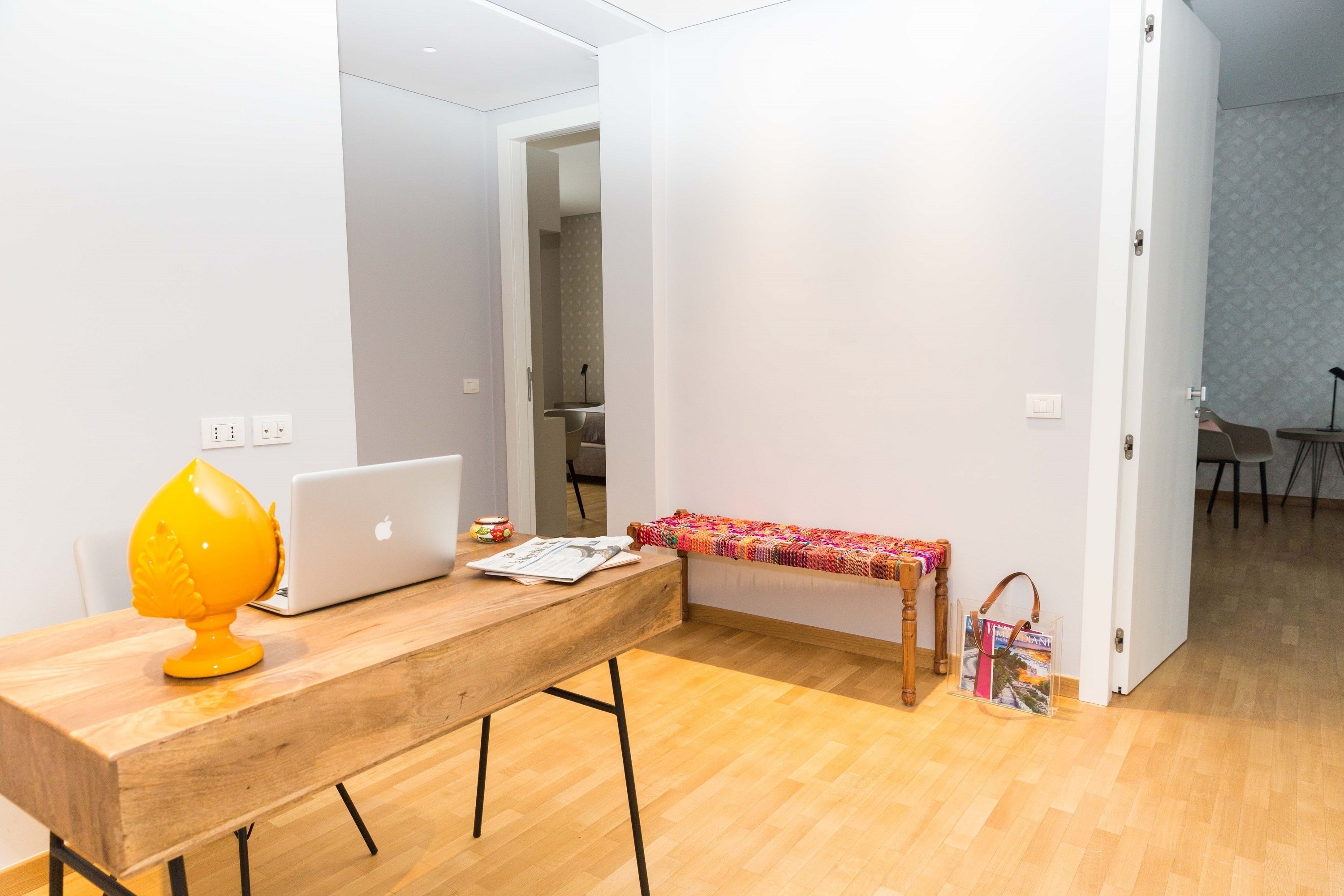 Una habitación con un escritorio de madera, una computadora portátil, una lámpara amarilla, una mesita con un mantel colorido y un bolso en el suelo.