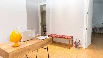 Una habitación con un escritorio de madera, una computadora portátil, una lámpara amarilla, una mesita con un mantel colorido y un bolso en el suelo.