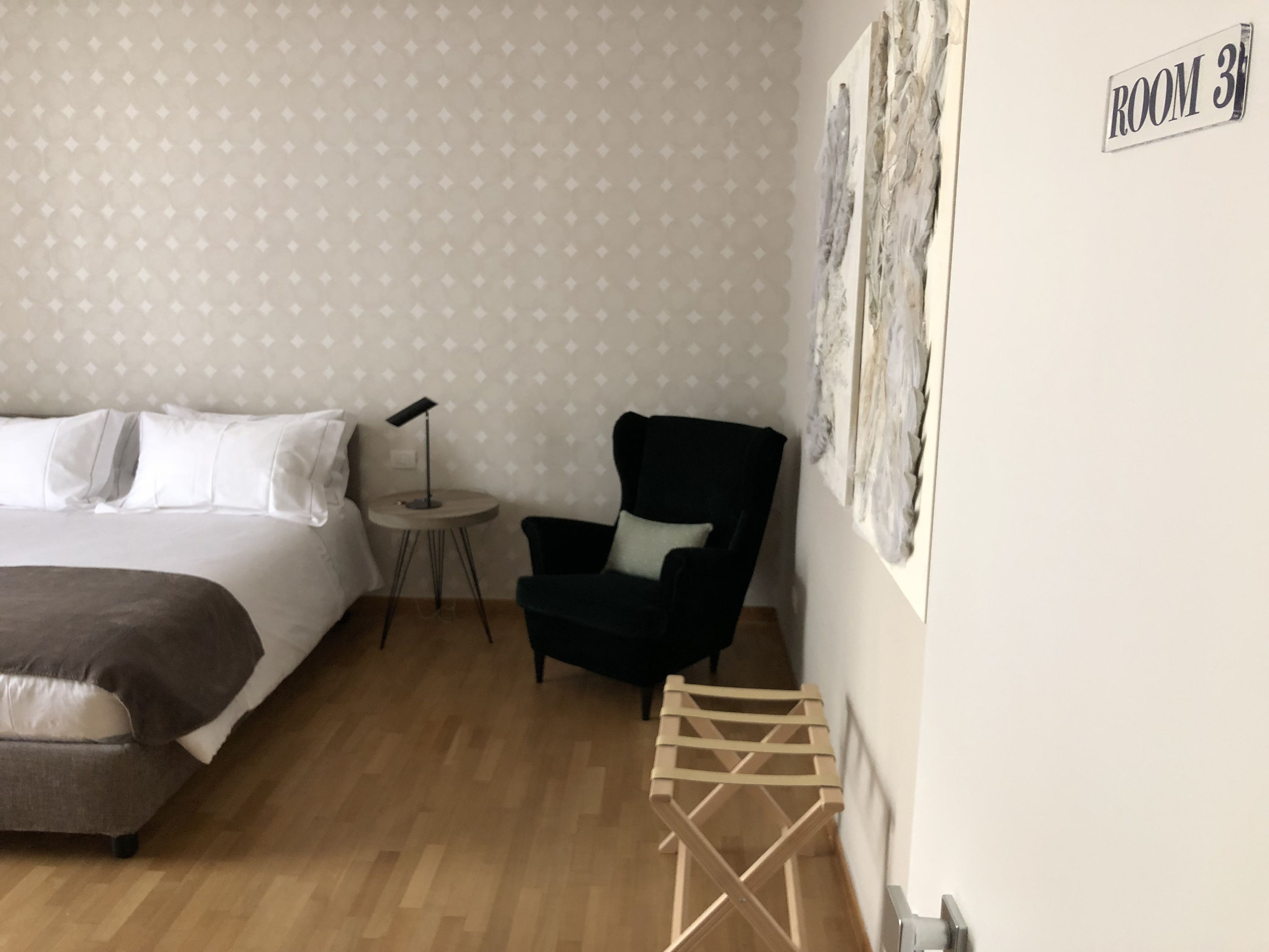 A hotel room with a bed, a small round table with a lamp, a dark armchair, and a wooden stool.
