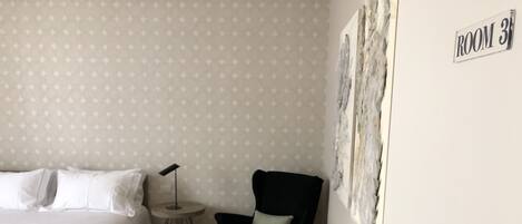 Una habitación de hotel con una cama, una mesita redonda con una lámpara, un sillón oscuro y un taburete de madera.