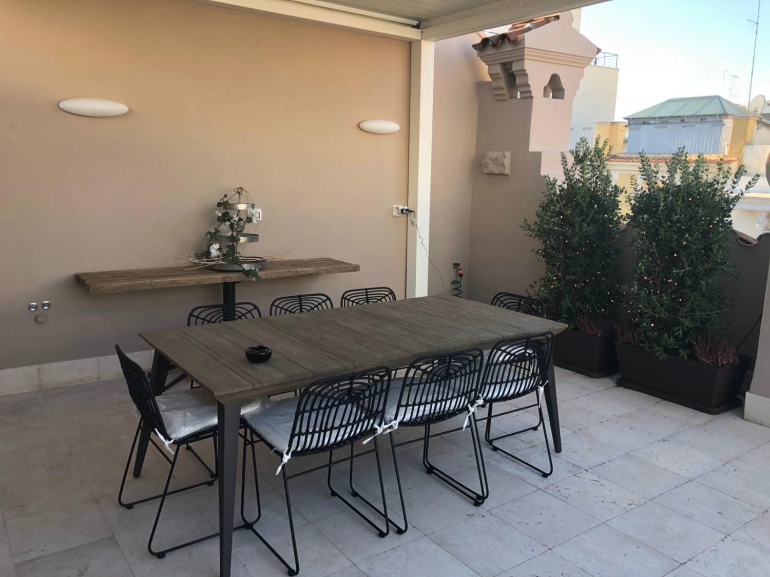 Un patio al aire libre con una mesa de madera y sillas de metal, rodeado por un muro color beige y plantas en macetas.