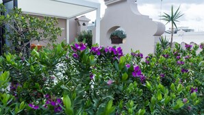 Un edificio moderno con fachada blanca, un arco distintivo y abundante vegetación con flores púrpuras.