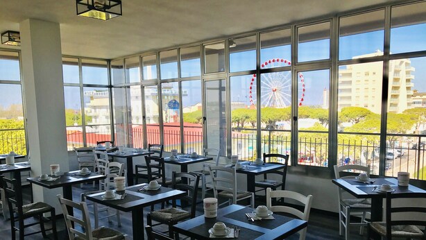 A restaurant with multiple tables and chairs, large windows offering a view of a Ferris wheel and buildings. - Hotel Cristina (Jesolo)