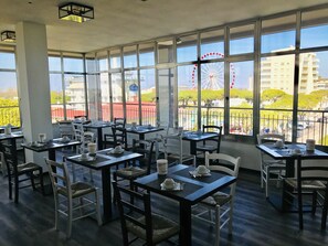 A restaurant with multiple tables and chairs, large windows offering a view of a Ferris wheel and buildings. - Hotel Cristina (Jesolo)
