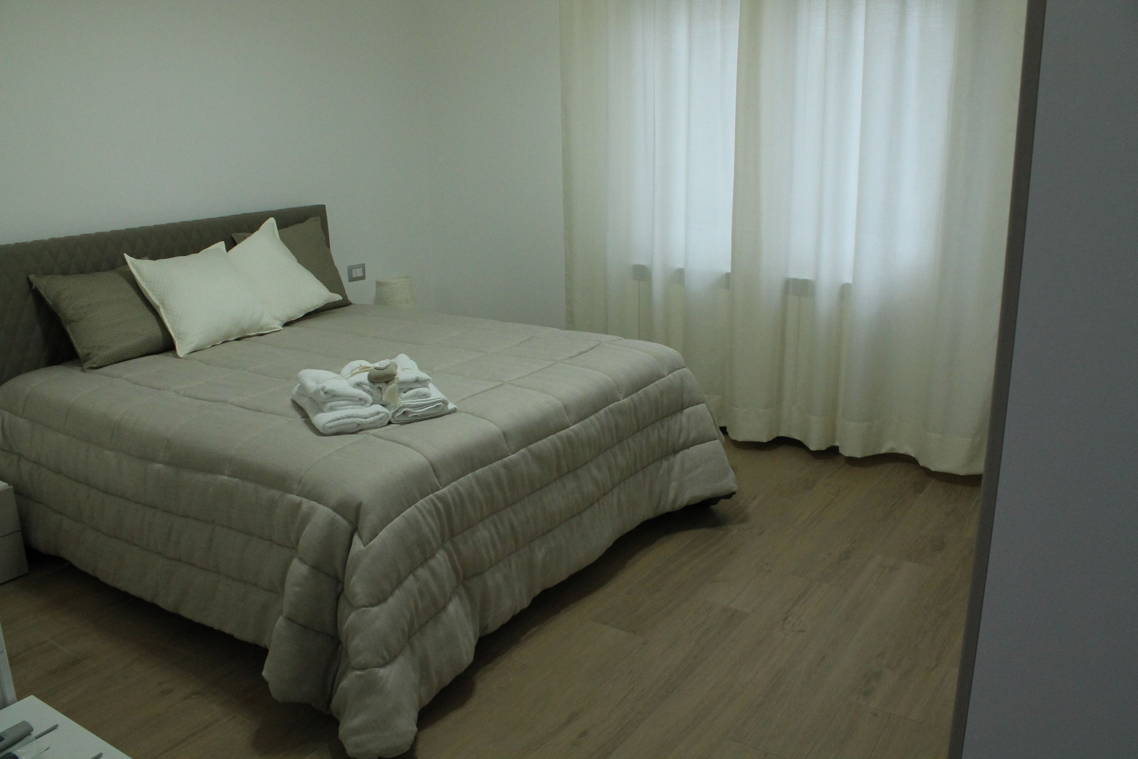 Ein ordentlich bezogenes Bett mit einer beigen Tagesdecke und Kissen, ein Holzboden und durchscheinende Vorhänge am Fenster.