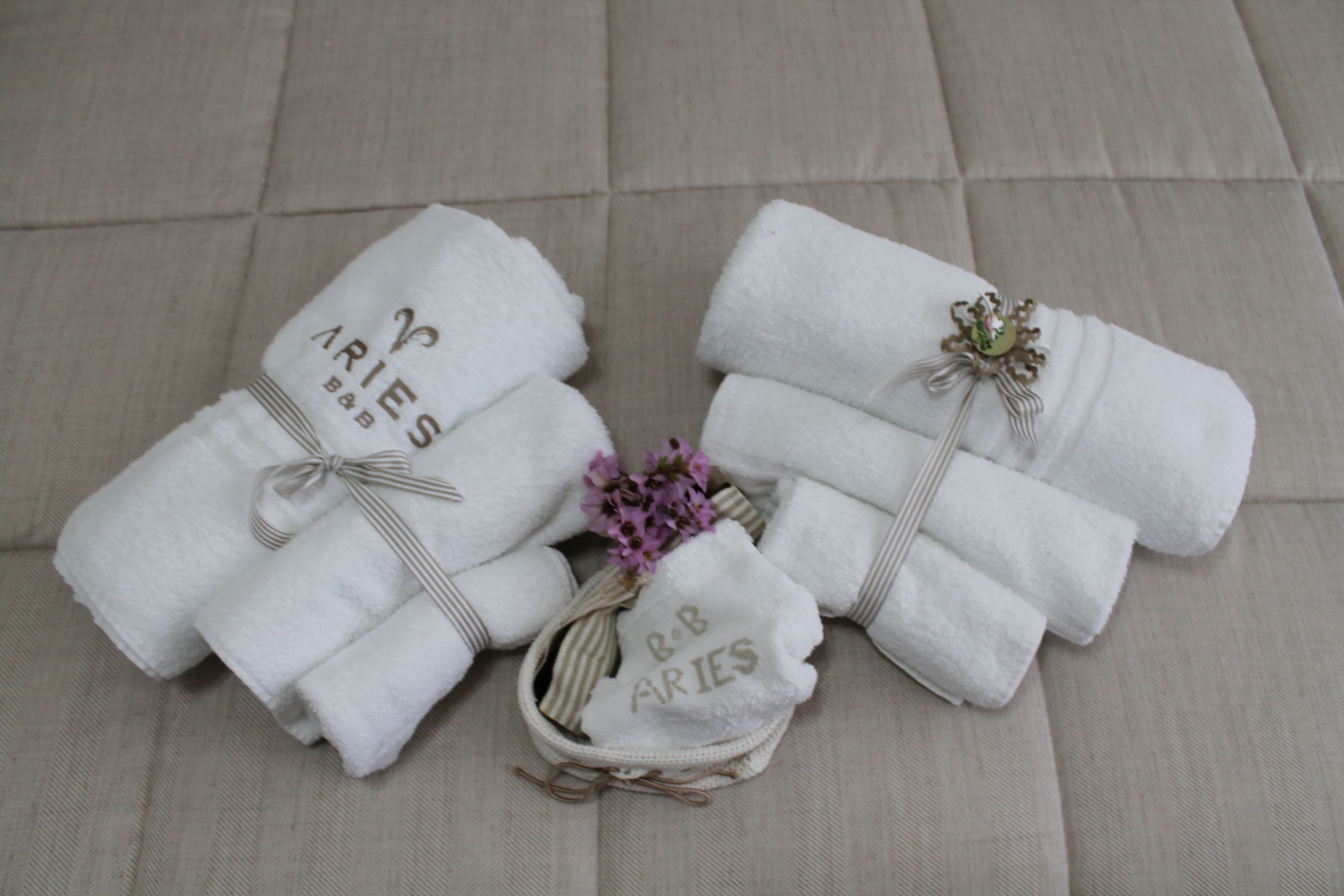 Two neatly folded white towels with "R&B ARIES" embroidered on them, tied with a ribbon, and placed on a beige sofa.
