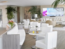 Un moderno vestíbulo de hotel con mobiliario blanco, una pantalla de televisión grande y plantas en macetas.