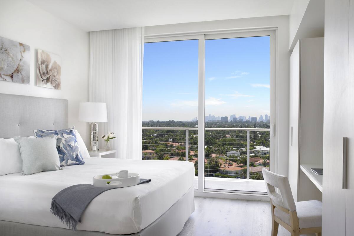 A modern hotel room with a large bed, a desk, and a balcony with a city view.