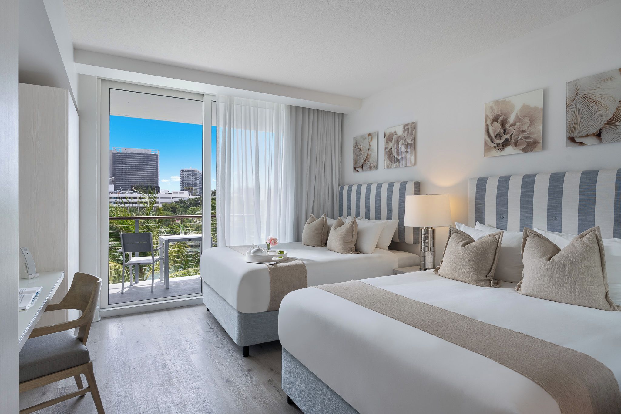 A hotel room with two beds, a desk, and a balcony with a city view.