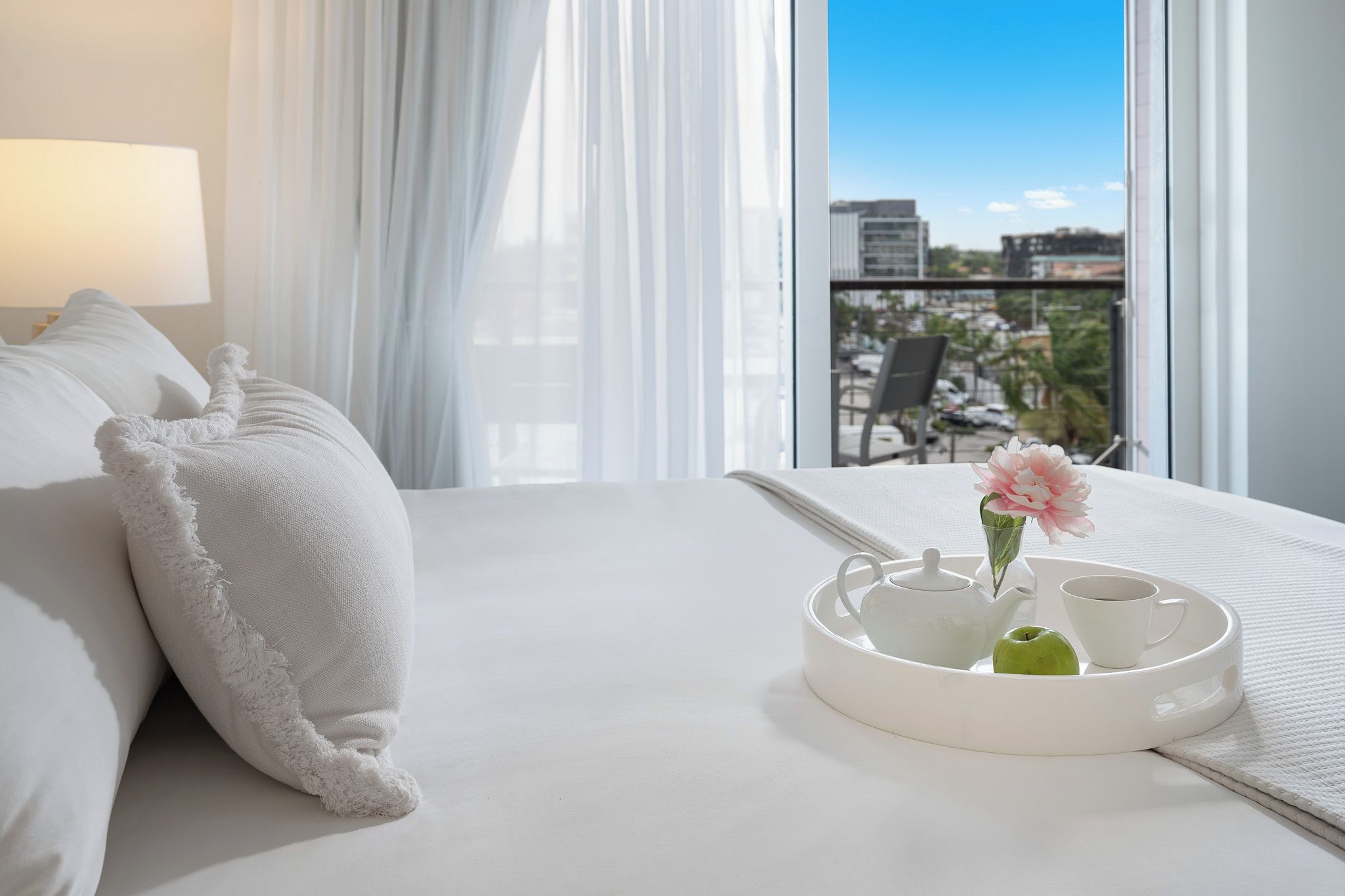 Een hotelkamer met een bed, een dienblad met een theepot, een kopje en een appel, en een uitzicht op de stad door het raam.