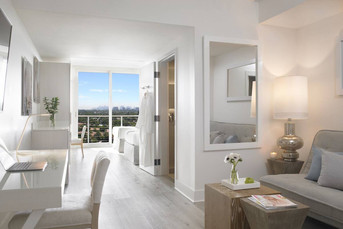 A modern hotel room with a balcony, a sitting area with a sofa and a table, and a workspace with a laptop.