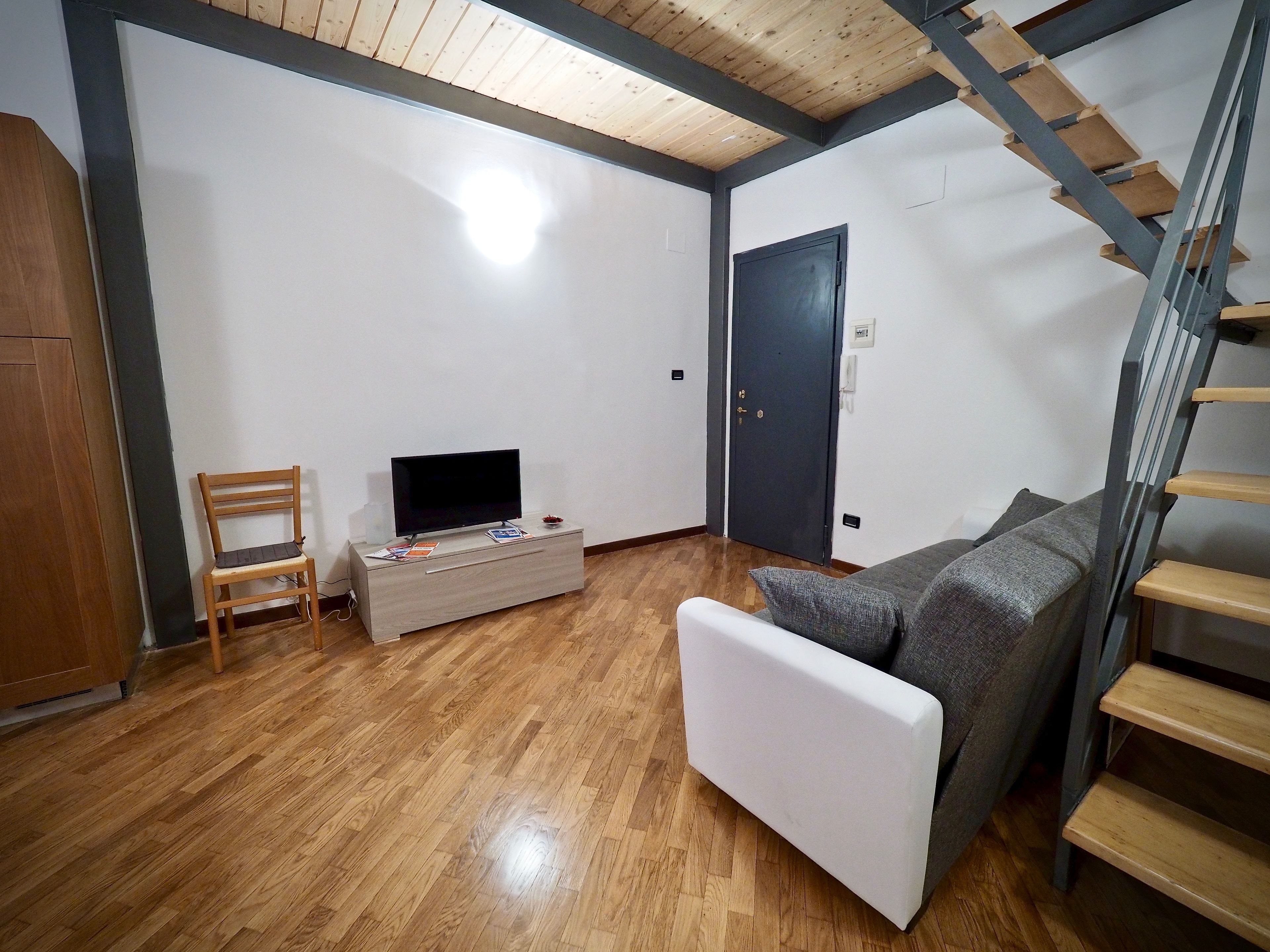 Uma sala de estar com piso de madeira, um sofá cinza, uma TV sobre um móvel de madeira, uma cadeira de madeira e uma escada.