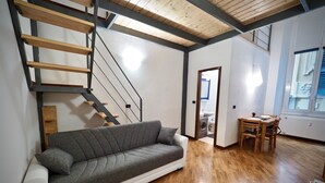 Un salon moderne avec un canapé gris, un plancher en bois, un escalier avec des rampes en métal et un coin repas comprenant une table et des chaises.
