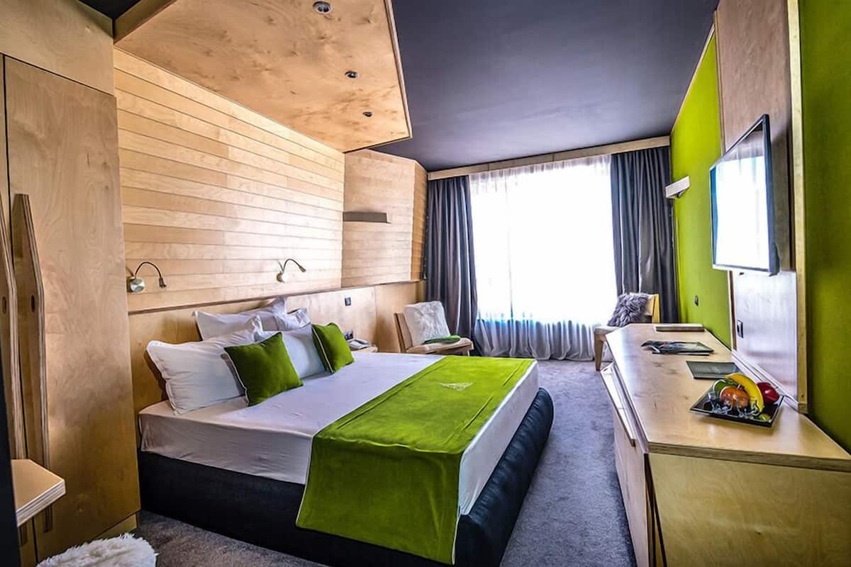 A modern hotel room with a wooden headboard, a bed with green and white bedding, a desk with a flat-screen TV, and a large window with curtains.