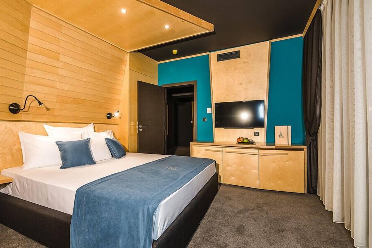 A modern hotel room with a wooden headboard, a bed with white and blue bedding, a desk with a flat-screen TV, and a teal accent wall.