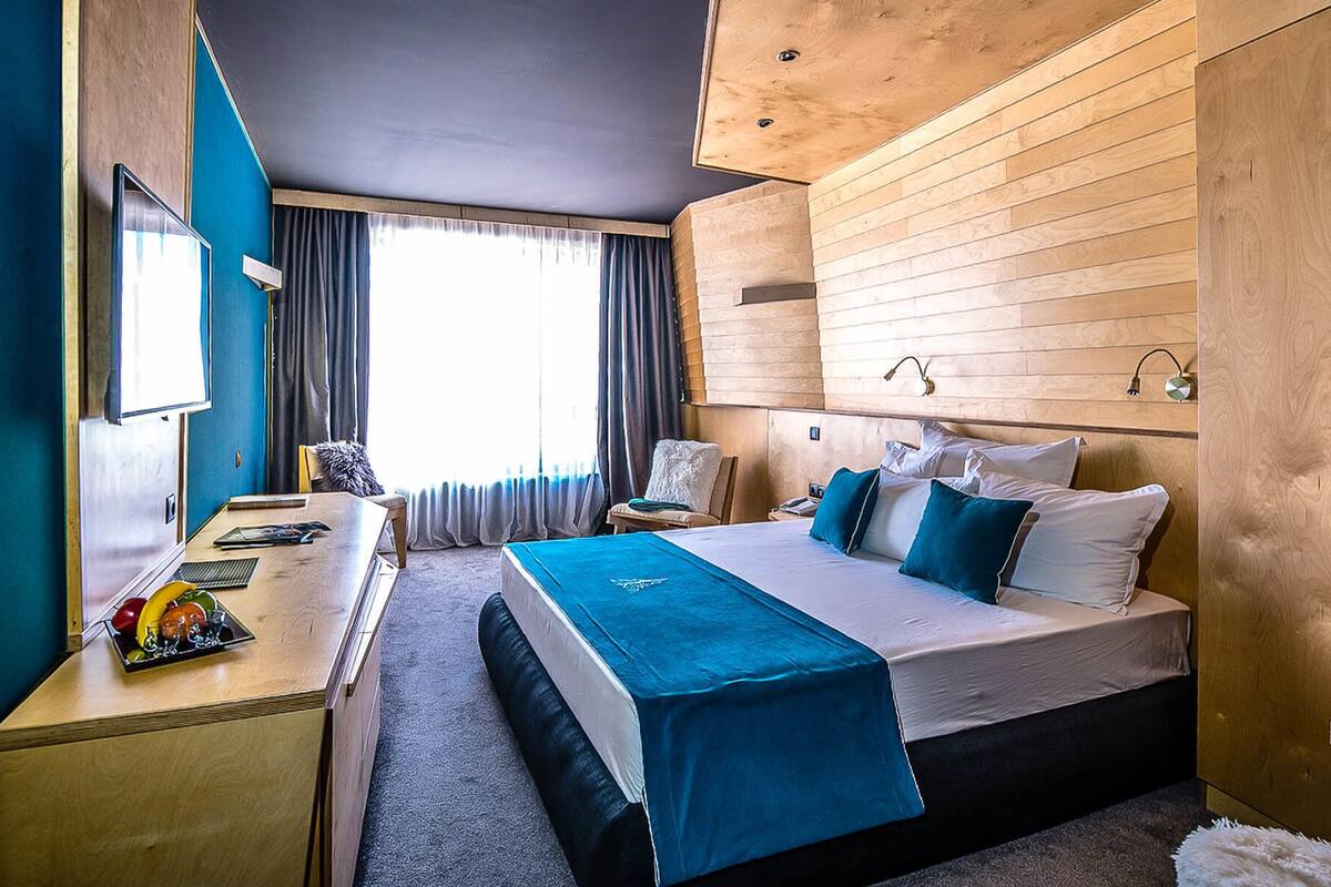A modern hotel room with a wooden headboard, a large bed with blue bedding, a desk with a computer, and a window with curtains.