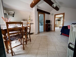 Amplia sala de estar con una mesa de comedor, sillas y un televisor. Se ve una cocina al fondo, y hay un radiador a la derecha.