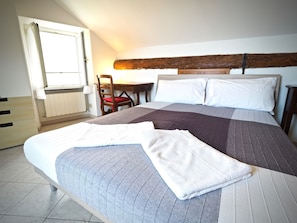 A neatly made bed with a striped blanket and pillows, a wooden chair with a red cushion, a wooden table, and a window with blinds. - Romantica Mansarda Pisano (Genoa)