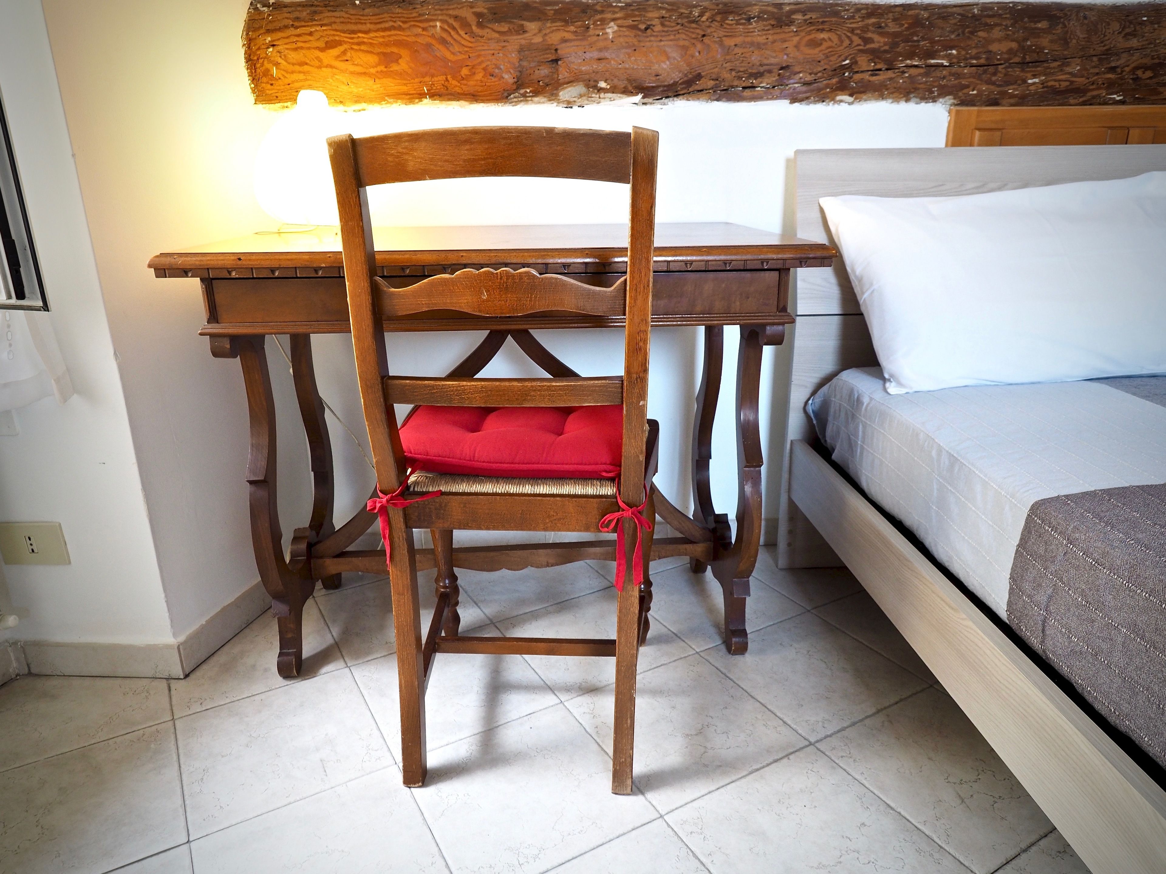 A wooden chair with a red cushion tied with a red ribbon, a wooden table, and a bed with white bedding.