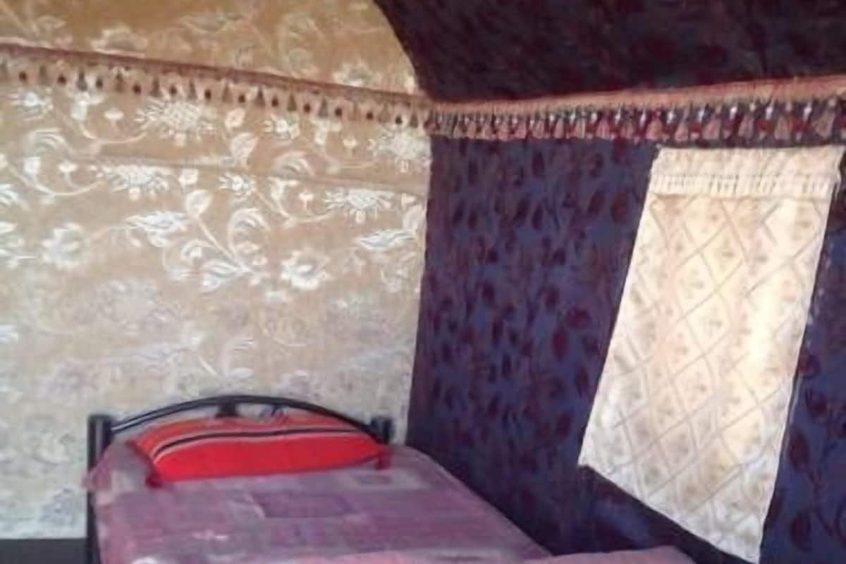 Uma cama de solteiro com colcha estampada e travesseiro vermelho em quarto com paredes padronizadas e janela com cortina.