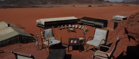 Un espacio al aire libre con sillas y mesas de mimbre, un brasero en el centro y tiendas de campaña en un paisaje desértico con formaciones rocosas al fondo.