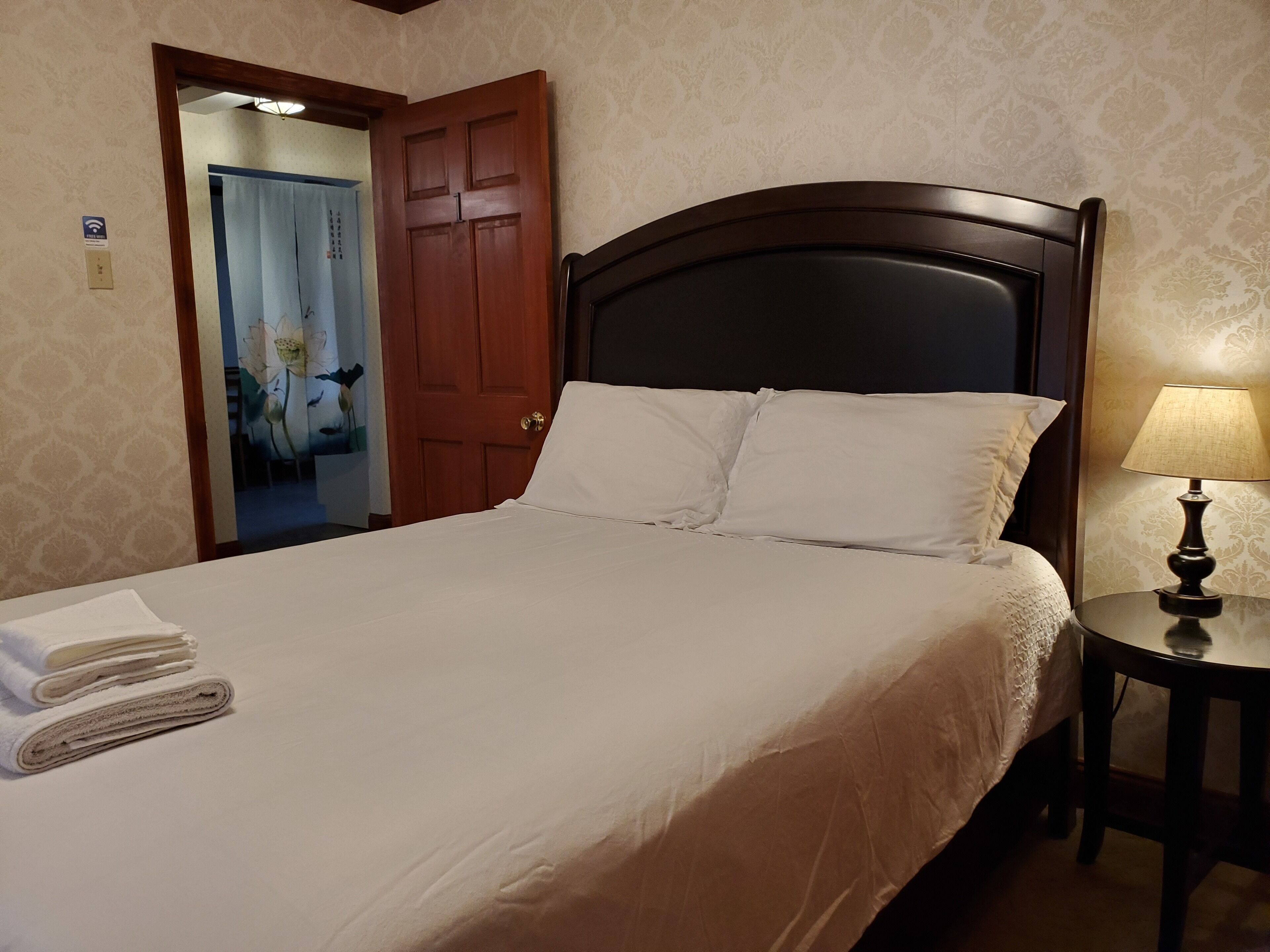 A neatly made bed with white linens and pillows, a wooden headboard, and a bedside table with a lamp.