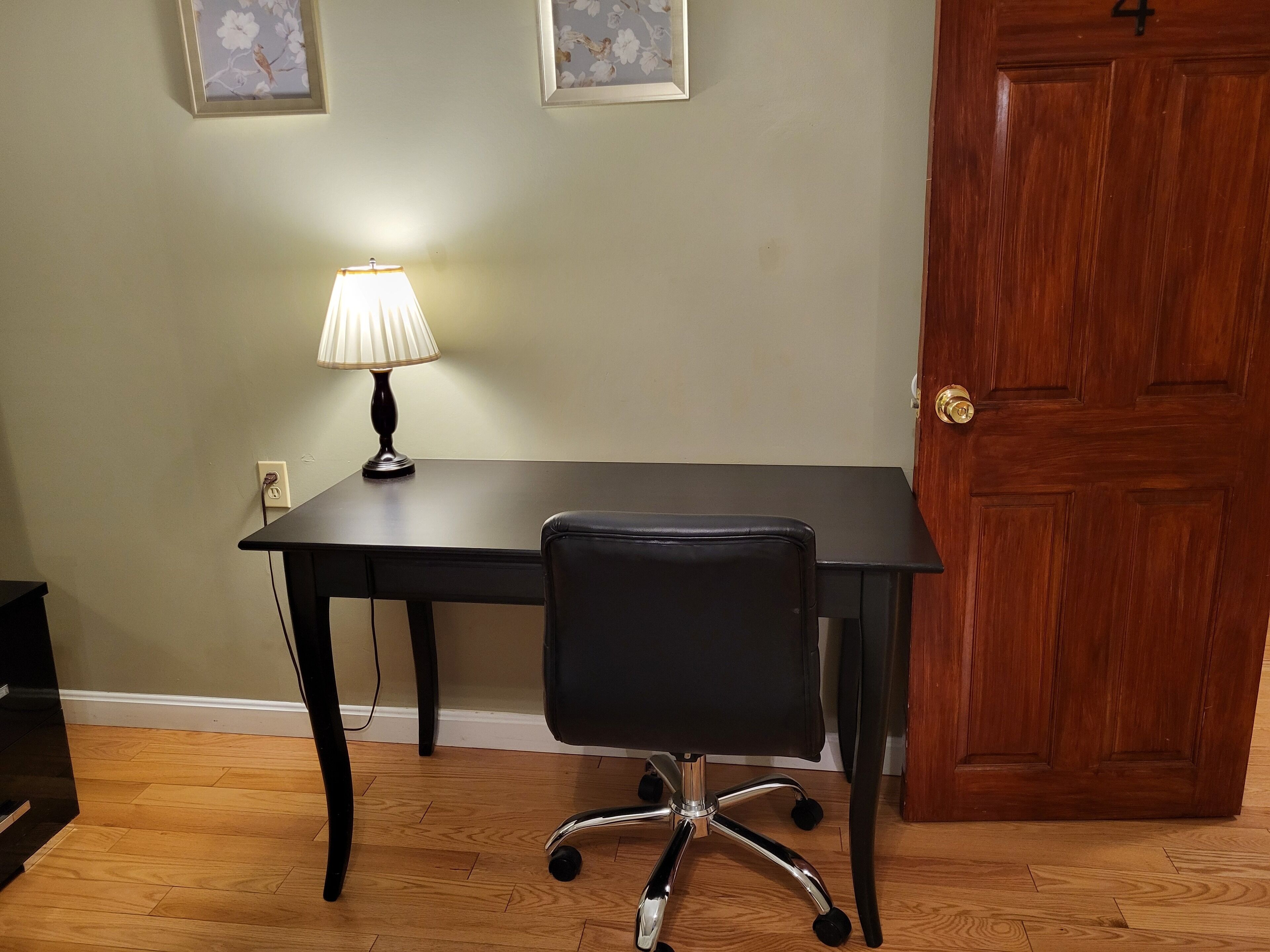 A room with a wooden floor, a dark wooden desk with a lamp, a black office chair, and a wooden door.