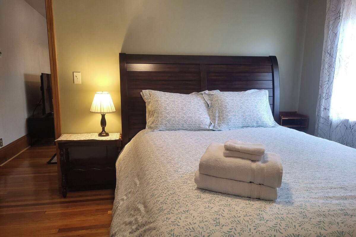 A neatly made bed with a floral-patterned bedspread, a bedside lamp, and a stack of folded towels on the bed.