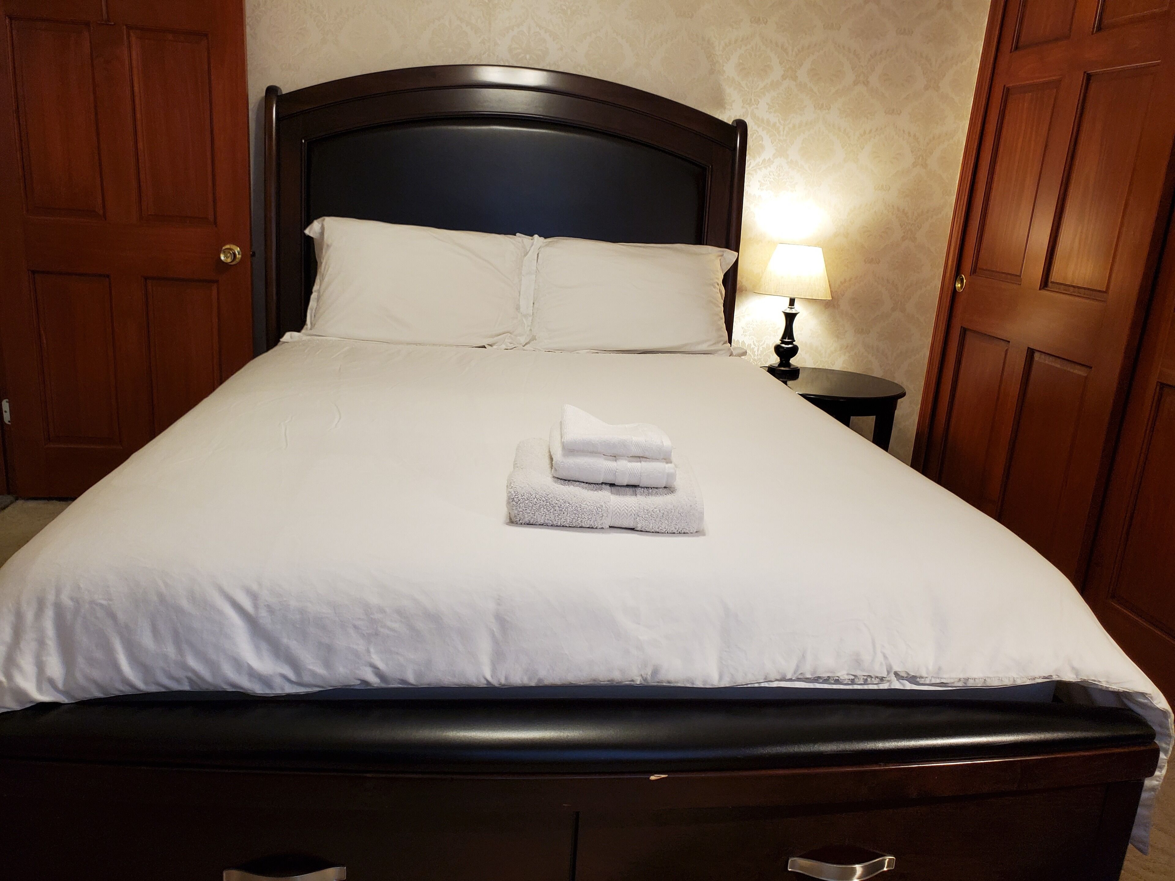 Una cama bien hecha con sábanas blancas y una toalla doblada, un cabecero de madera y una lámpara de noche.