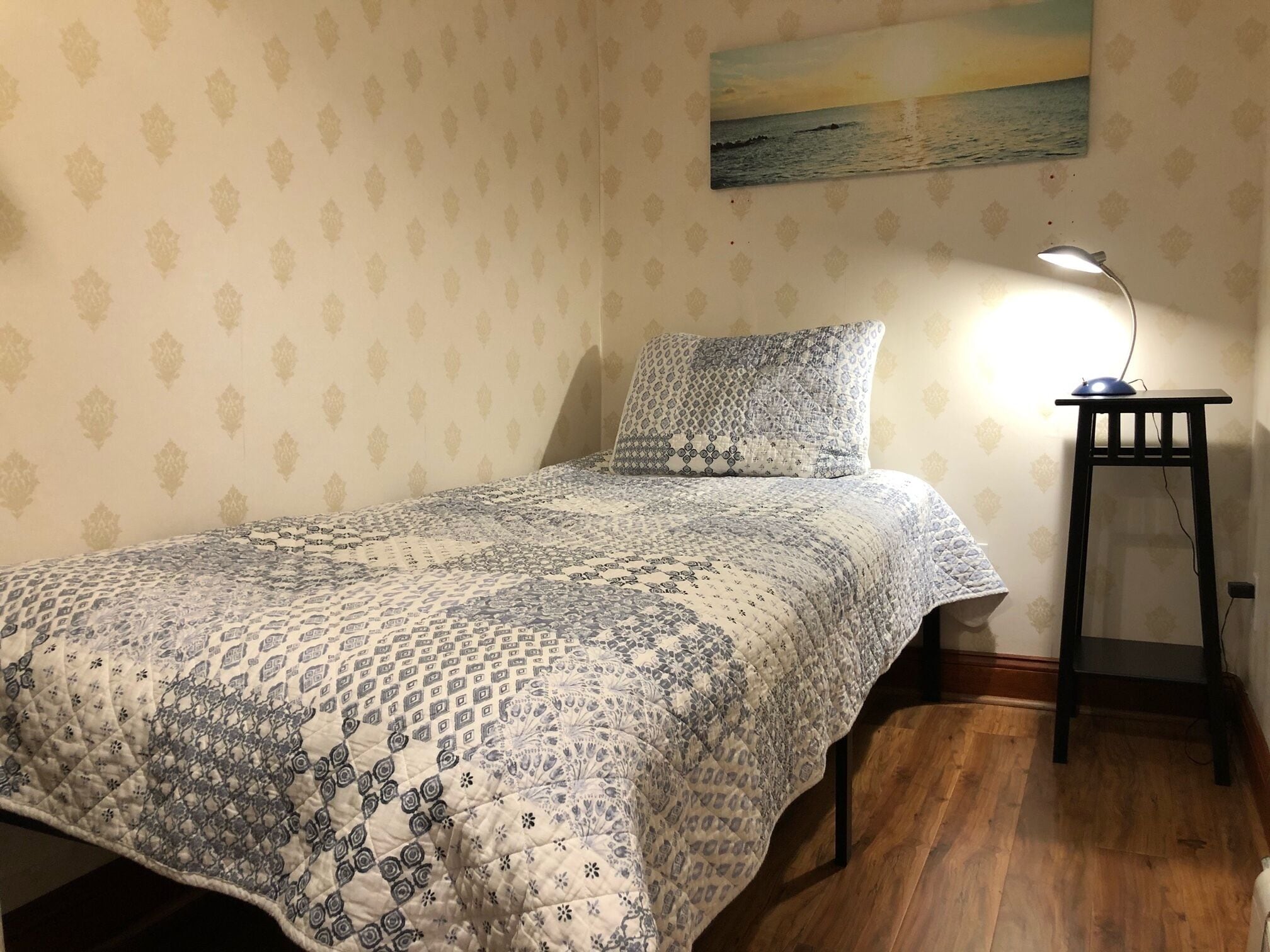 A bedroom with a patterned bedspread, a pillow, a nightstand with a lamp, and a painting of a sunset on the wall.