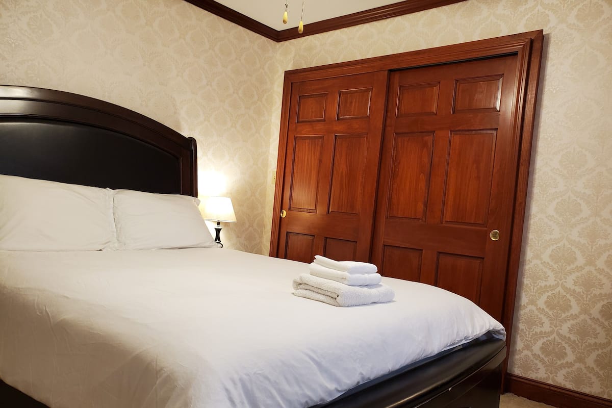 A neatly made bed with white linens and a folded towel, a wooden nightstand with a lamp, and a large wooden door with a brass doorknob.