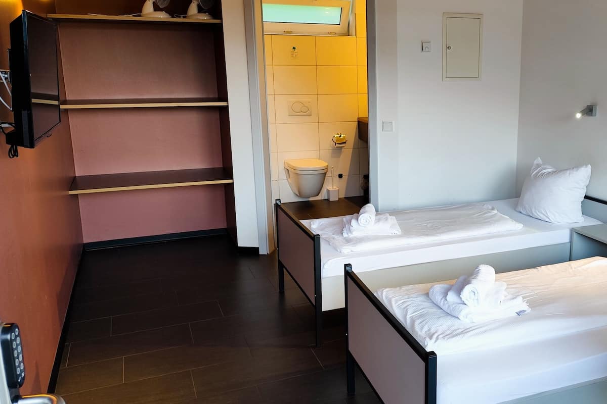 Une chambre d’hôtel avec deux lits simples, un petit bureau, une chaise, un téléviseur fixé au mur et une salle de bain avec des toilettes et un lavabo.