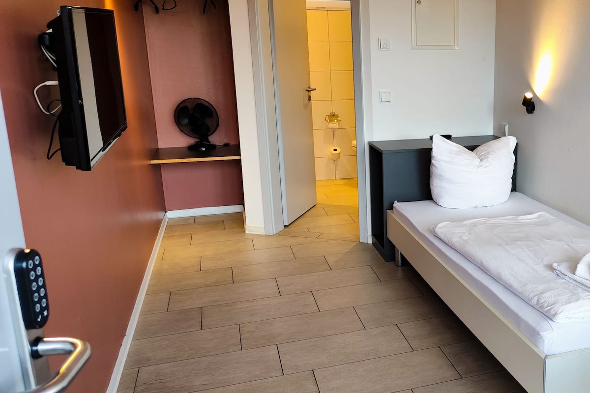 Une chambre d’hôtel avec un lit simple, une table de chevet, un petit bureau, un téléviseur fixé au mur et une salle de bain visible à travers une porte ouverte.