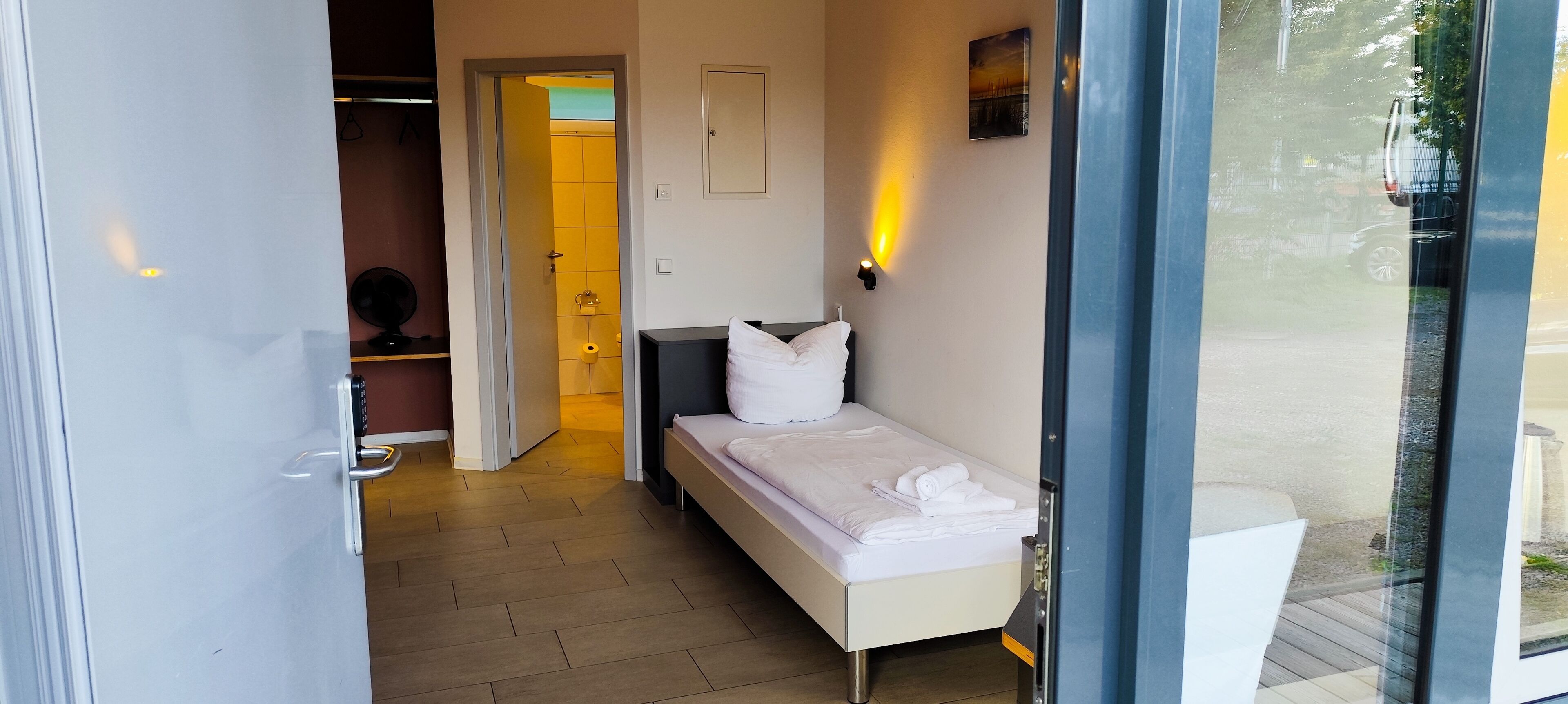 Una habitación de hotel con una cama, una mesita de noche, una lámpara de pared y una puerta que da al baño.