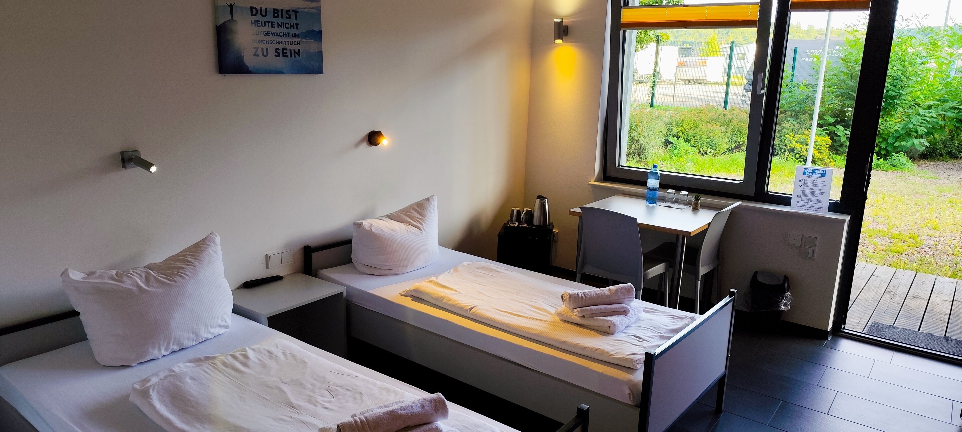 A hotel room with two beds, a desk, and a window with a view of greenery.