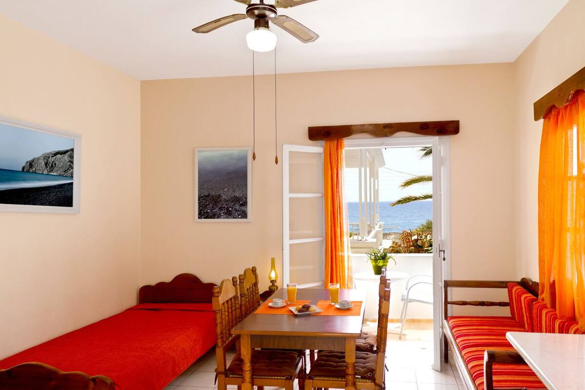 A room with a bed, a dining table with chairs, a ceiling fan, and a view of the sea through an open door.